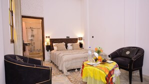 A hotel room with a bed, a table with a water bottle and fruit, two chairs, and a shower area. - RYAD RDATE LWALIDINE (Marrakech)