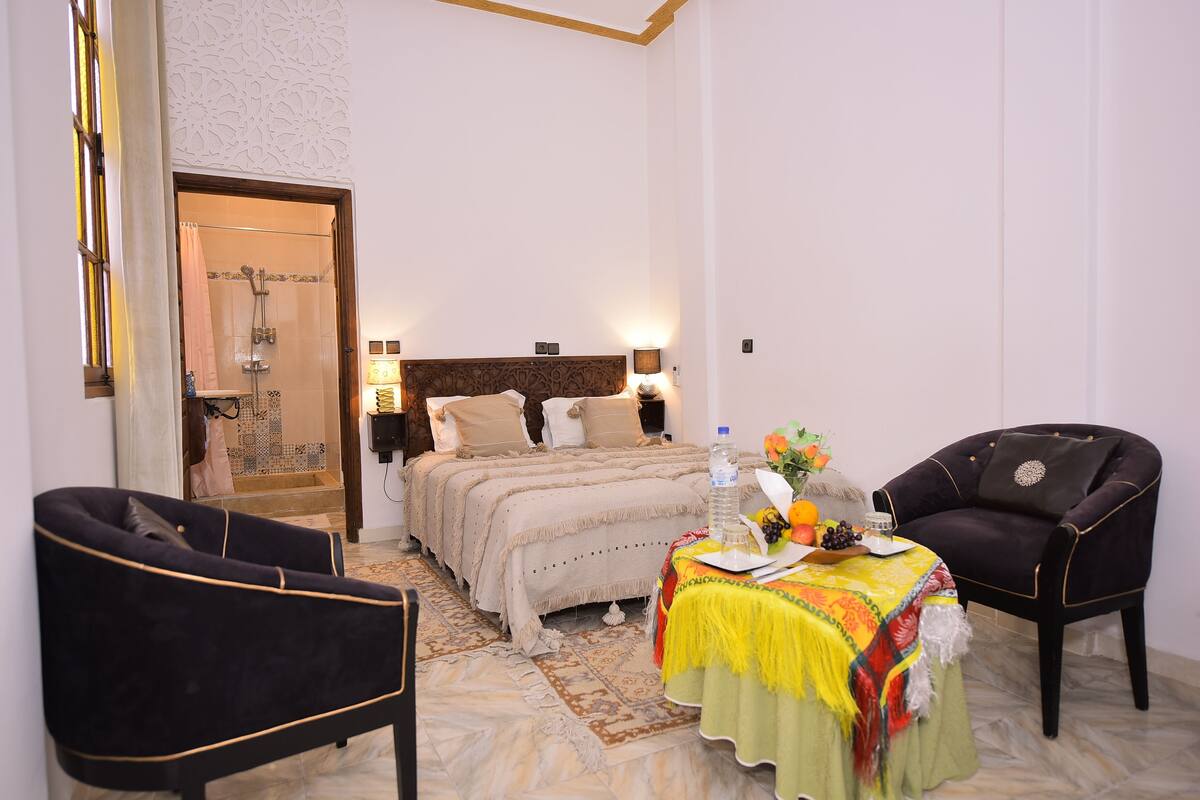 A hotel room with a bed, a table with a water bottle and fruit, two chairs, and a shower area.