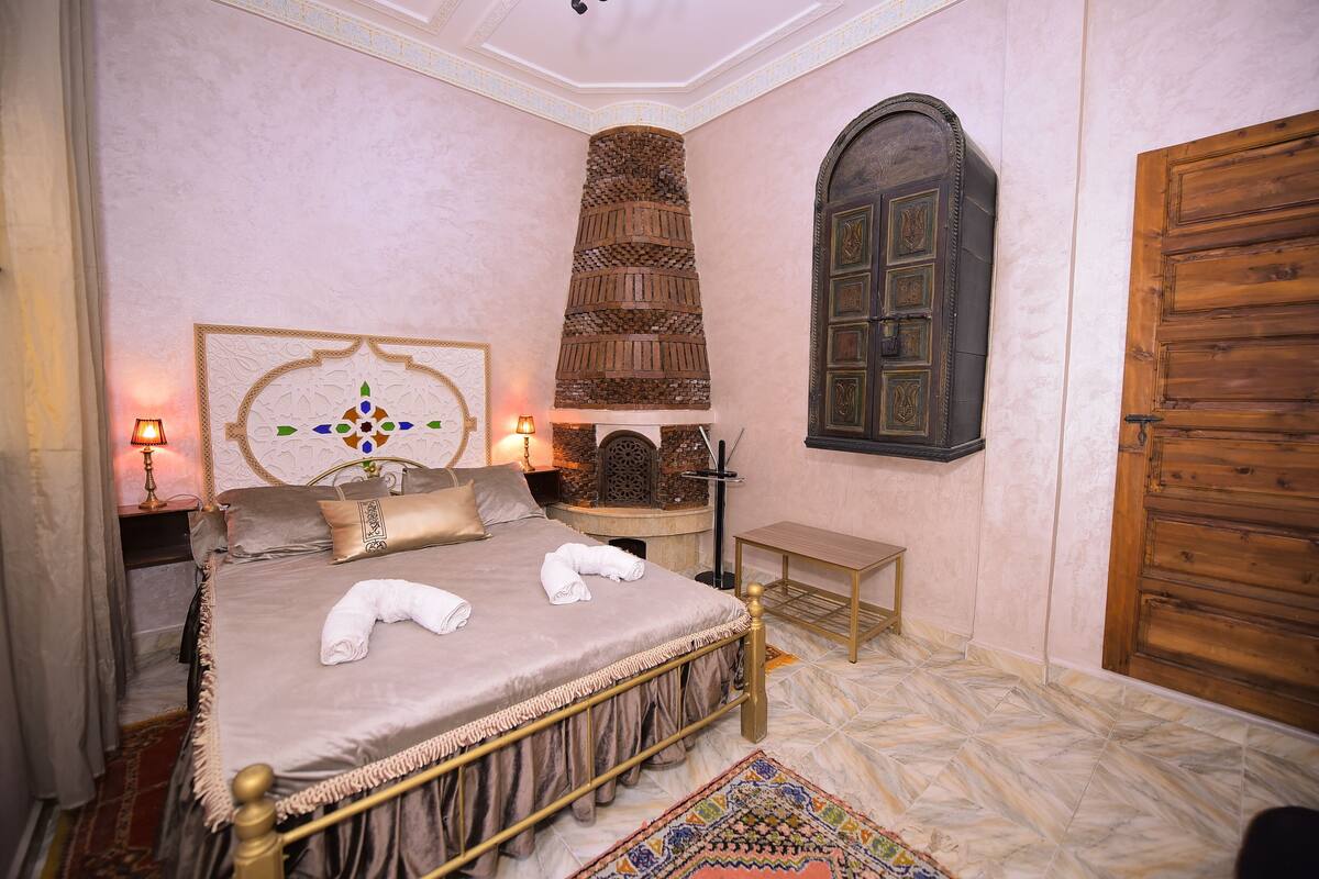 A neatly arranged bedroom with a bed, bedside lamps, a decorative headboard, a wooden chest, and a large wooden door.