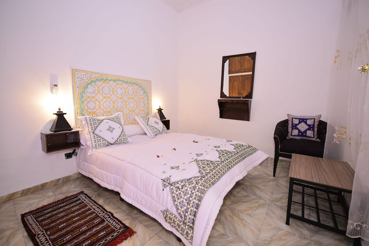A bedroom with a bed, nightstands, a wooden chair, a small table, and a wall-mounted shelf.