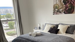 A modern bedroom with a large bed, a bedside table, a nightstand, and a view of the street through the window.