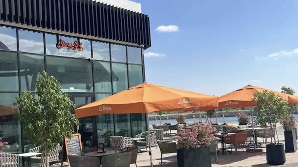 An outdoor seating area with tables and chairs under orange umbrellas, in front of a glass building with a sign.