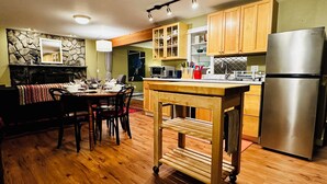 A kitchen with wooden cabinets, a stainless steel refrigerator, a dining table with chairs, and a stone wall.