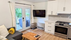 A compact living space with a sofa, a coffee table, a TV mounted on the wall, a kitchen area with a stove and oven, and a sliding glass door leading to an outdoor area. - Private Garden Suite in Langford - Free car charger (Langford)