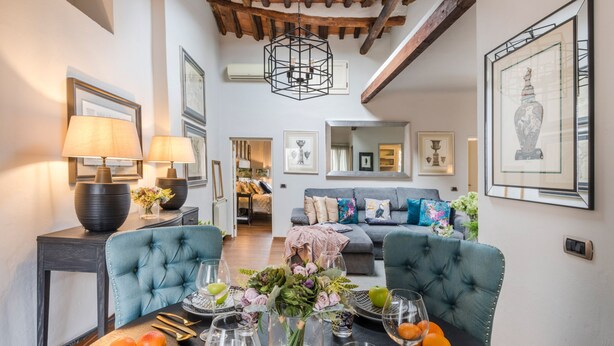 A dining area with a table set for four, featuring a vase of flowers, wine glasses, and fruit. There are two blue upholstered chairs, a dark side table with lamps, and a sofa in the background. - Clocktower Penthouse (Lucca)