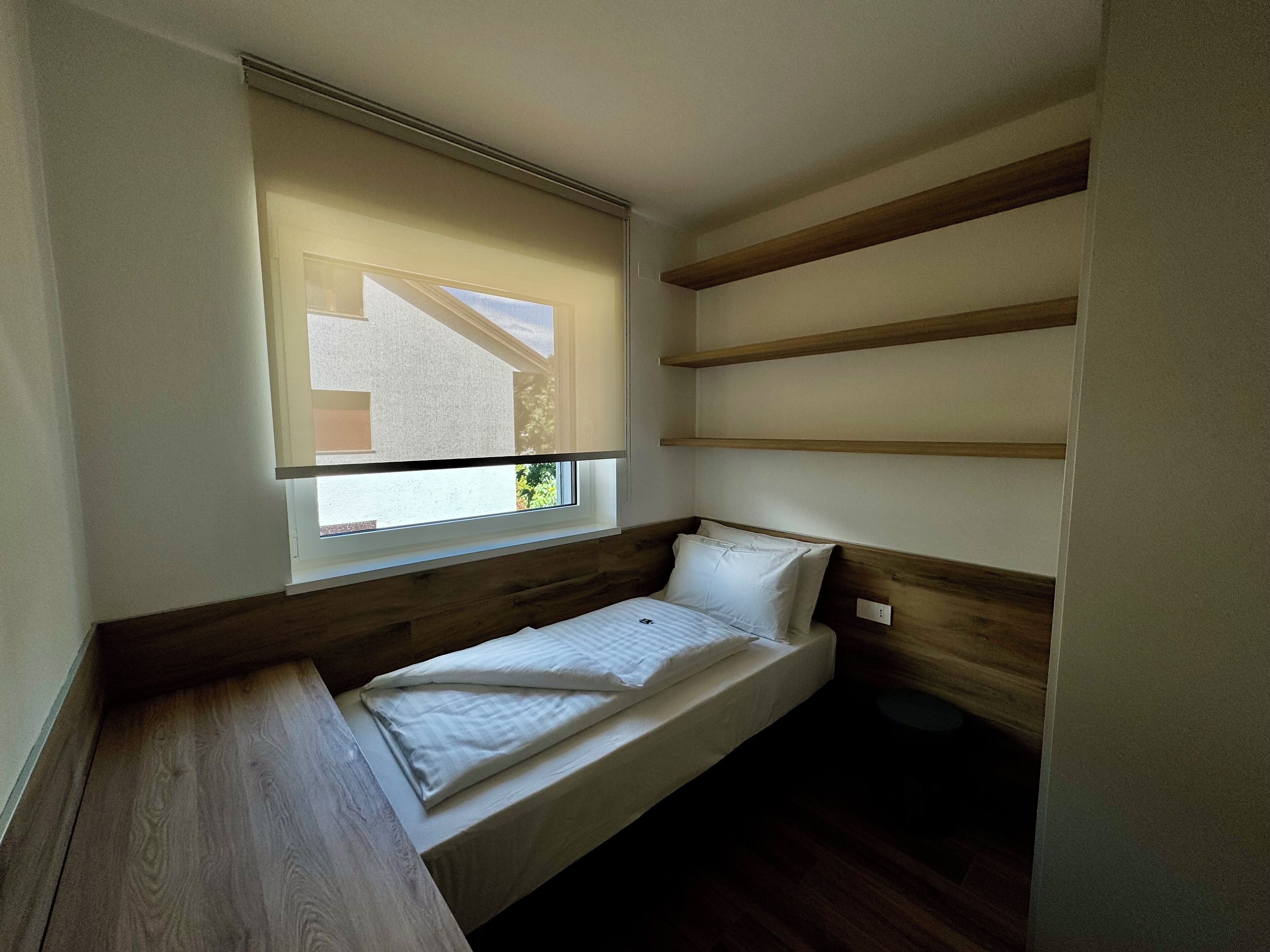 A small, single-bed room with a wooden headboard, a window with blinds, and built-in shelves.