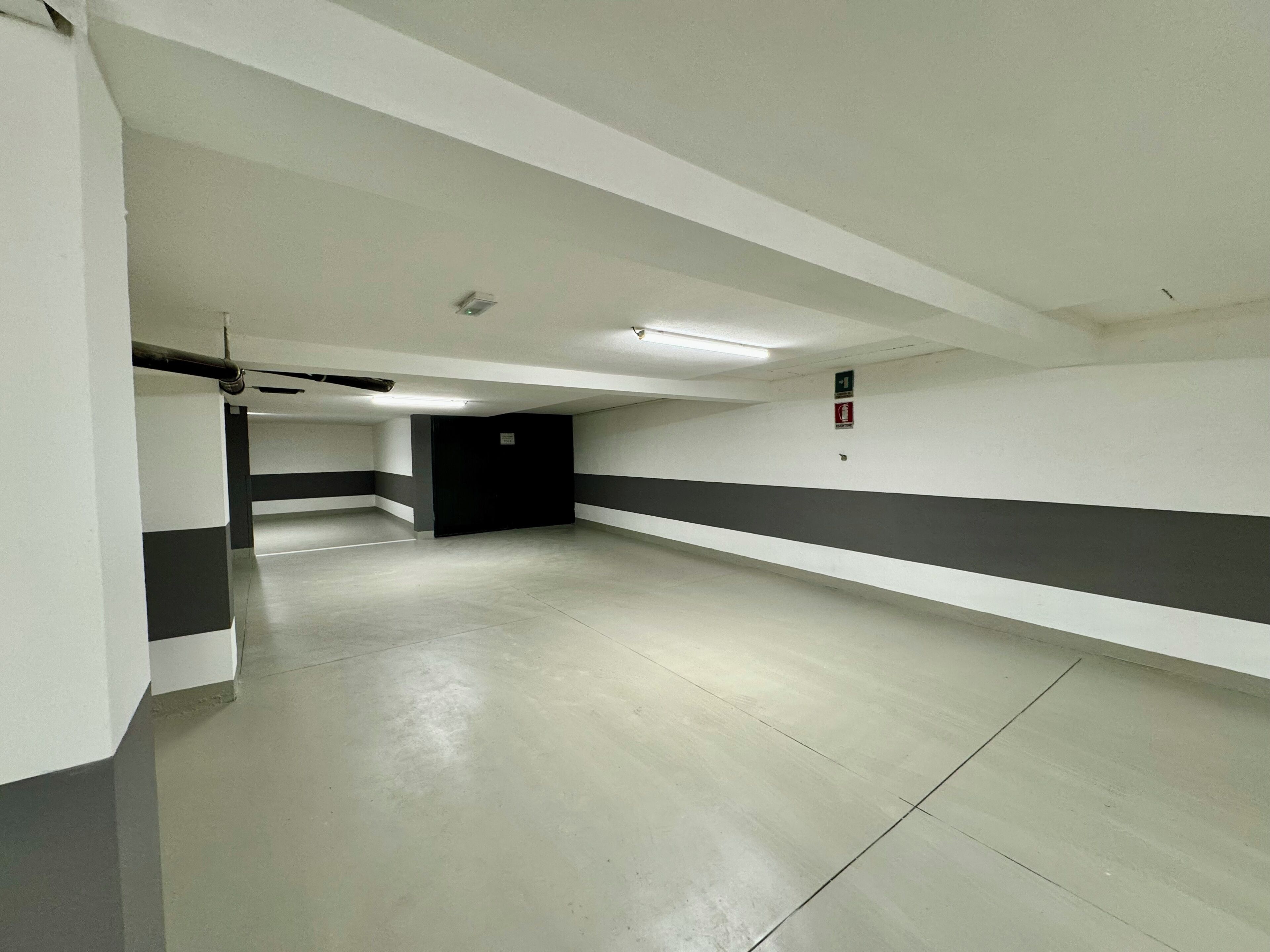 Un long couloir spacieux et vide, aux murs blancs et gris, doté d’un sol en béton et d’une porte noire au bout.