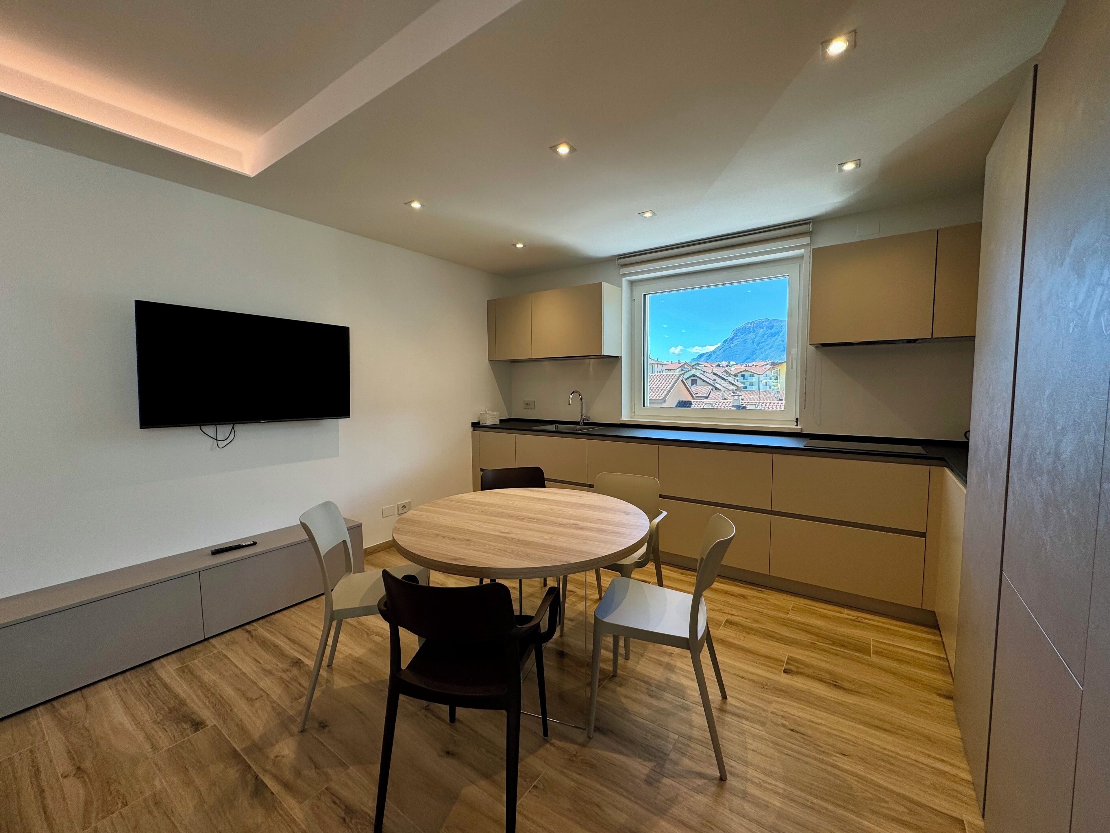 A modern kitchen with a round dining table, chairs, a flat-screen TV, and a window with a mountain view.
