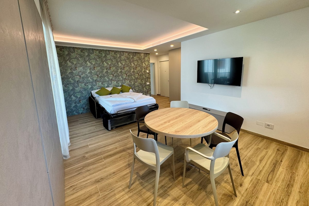 A modern hotel room with a bed, a TV, a round dining table with chairs, and a wooden floor.
