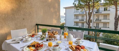 A breakfast table set with eggs, bacon, toast, and juice on a balcony overlooking a building and trees.