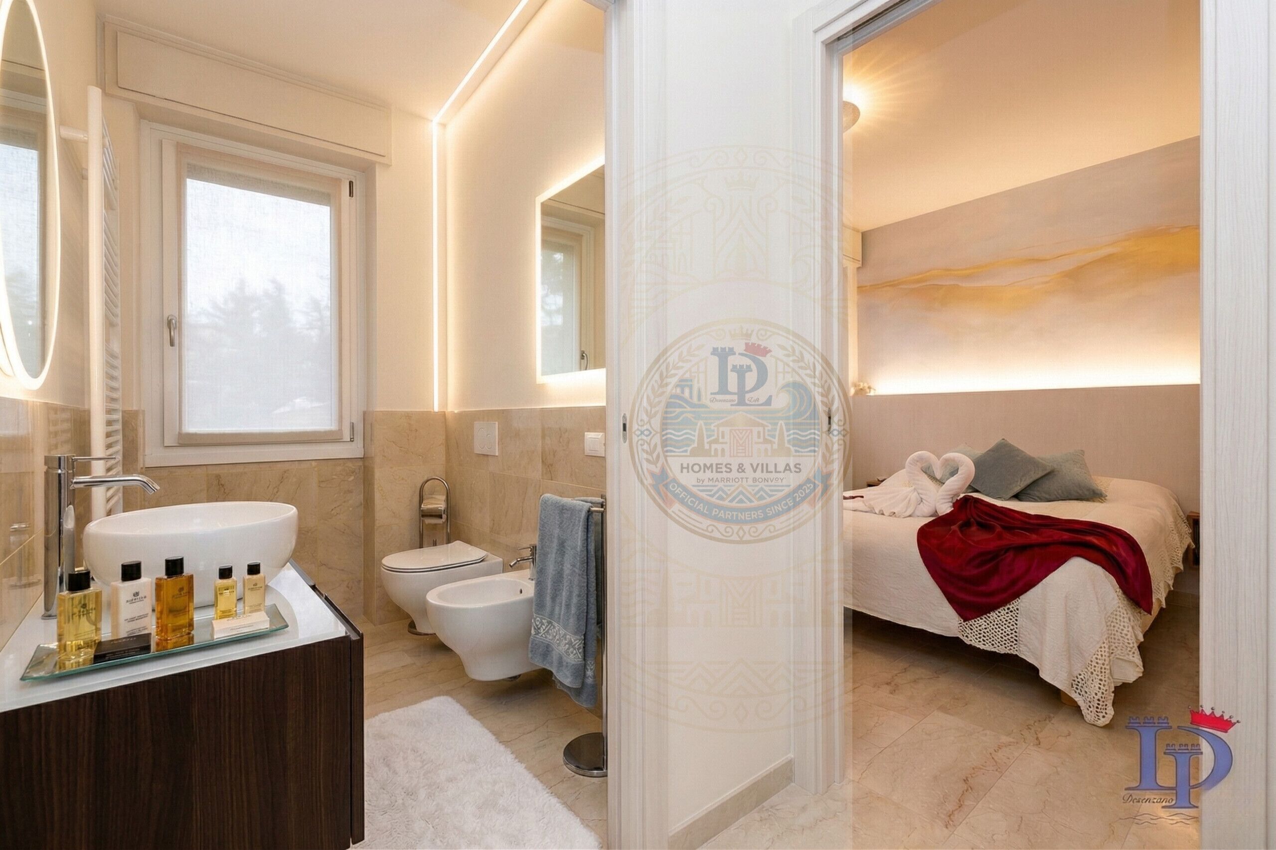 A modern bathroom with a large sink, toilet, and bidet. There is a bed with a red blanket and pillows.
