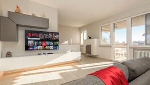 A modern living room with a flat-screen TV mounted on a white console, a grey sofa with a red throw, and a balcony with a view.