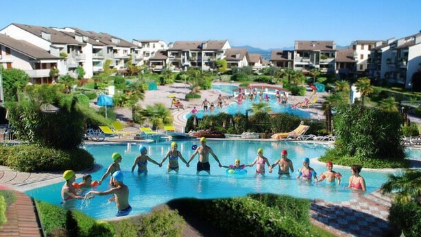 A swimming pool area with people holding hands, surrounded by greenery and modern buildings.