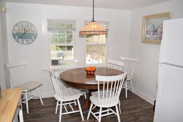 A dining area with a round wooden table surrounded by white chairs, a hanging light fixture, and a clock on the wall. - Surf & Wind- Fire Pit, Fenced in Yard (Chincoteague)