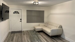 A living room with a beige sofa, a glass coffee table, a door with a decorative glass window, and a ceiling light fixture. - Canal Home with Pool near Beaches and Space Center! (Merritt Island)