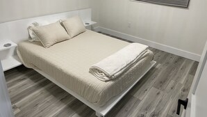 A neatly made bed with a white headboard and footboard, a bedside table, and a wall-mounted window with blinds. - Canal Home with Pool near Beaches and Space Center! (Merritt Island)