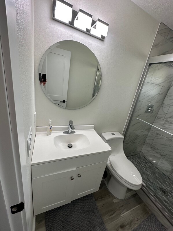 A modern bathroom with a white vanity, a round mirror, a toilet, and a glass shower enclosure.