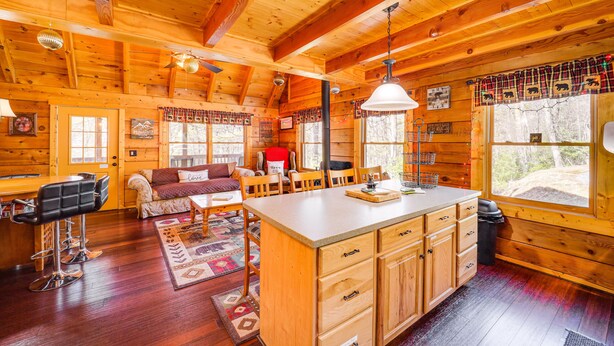 A cozy cabin interior with a wooden interior, a dining area, a kitchen with wooden cabinets, and a large window providing a view of the outdoors. - Cozy Family Cabin w/ Hot Tub - 10 Mi to App State! (Deep Gap)