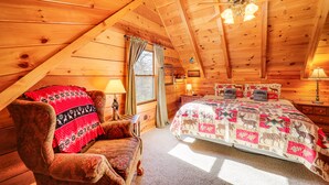 A cozy log cabin bedroom with a wooden ceiling, a bed with a patterned quilt, a chair with a red blanket, and a window with curtains. - Cozy Family Cabin w/ Hot Tub - 10 Mi to App State! (Deep Gap)