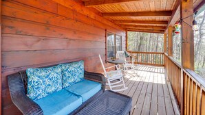 A wooden deck with a wicker sofa and rocking chairs. - Cozy Family Cabin w/ Hot Tub - 10 Mi to App State! (Deep Gap)