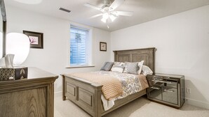 Une chambre à coucher avec un lit en bois, une table de chevet, une commode, un ventilateur de plafond et une fenêtre avec des stores.