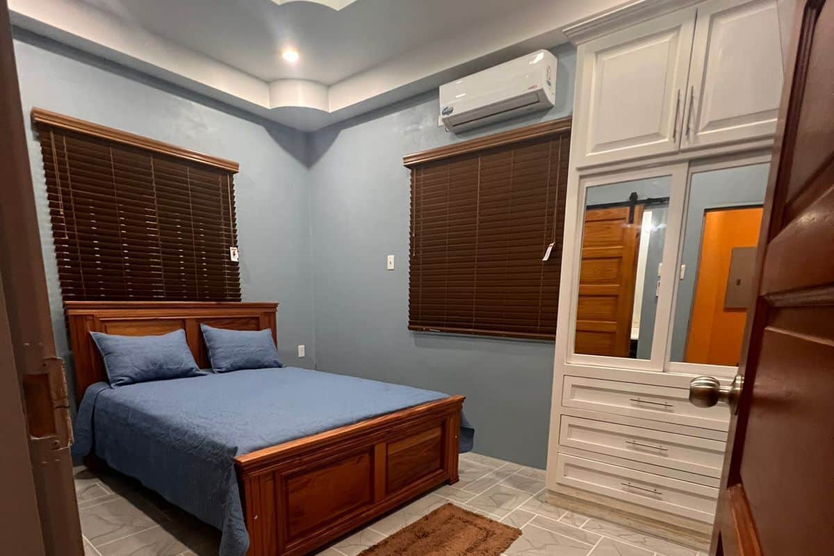 A bedroom with a bed, wooden headboard, two windows with blinds, an air conditioning unit, a brown rug, and a white dresser.
