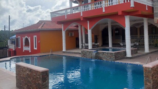 A large swimming pool with a stone border, a red and yellow building with a balcony, and a cloudy sky. - Aqua Lounge The Venue (Sangre Grande)