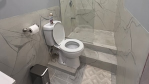 A bathroom with a toilet, a glass-enclosed shower, and a marble-patterned wall. - Aqua Lounge The Venue (Sangre Grande)
