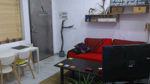Una habitación pequeña y amoblada con un sofá rojo, un escritorio negro, una computadora, una mesita y una planta en maceta.