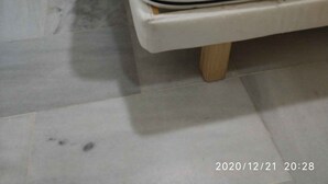 Una cama con colchón blanco y estructura de madera, un taburete de madera pequeño, y piso de baldosas.