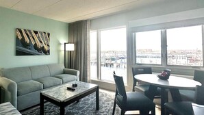 Une chambre d’hôtel comprenant un canapé, une table basse, une table à manger avec des chaises, une grande fenêtre donnant sur un port de plaisance et un tableau accroché au mur.