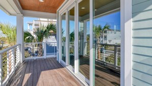 A covered wooden deck with glass doors leading to a balcony, overlooking a residential area with houses and palm trees. - Good Vibes Villa (St. Augustine)