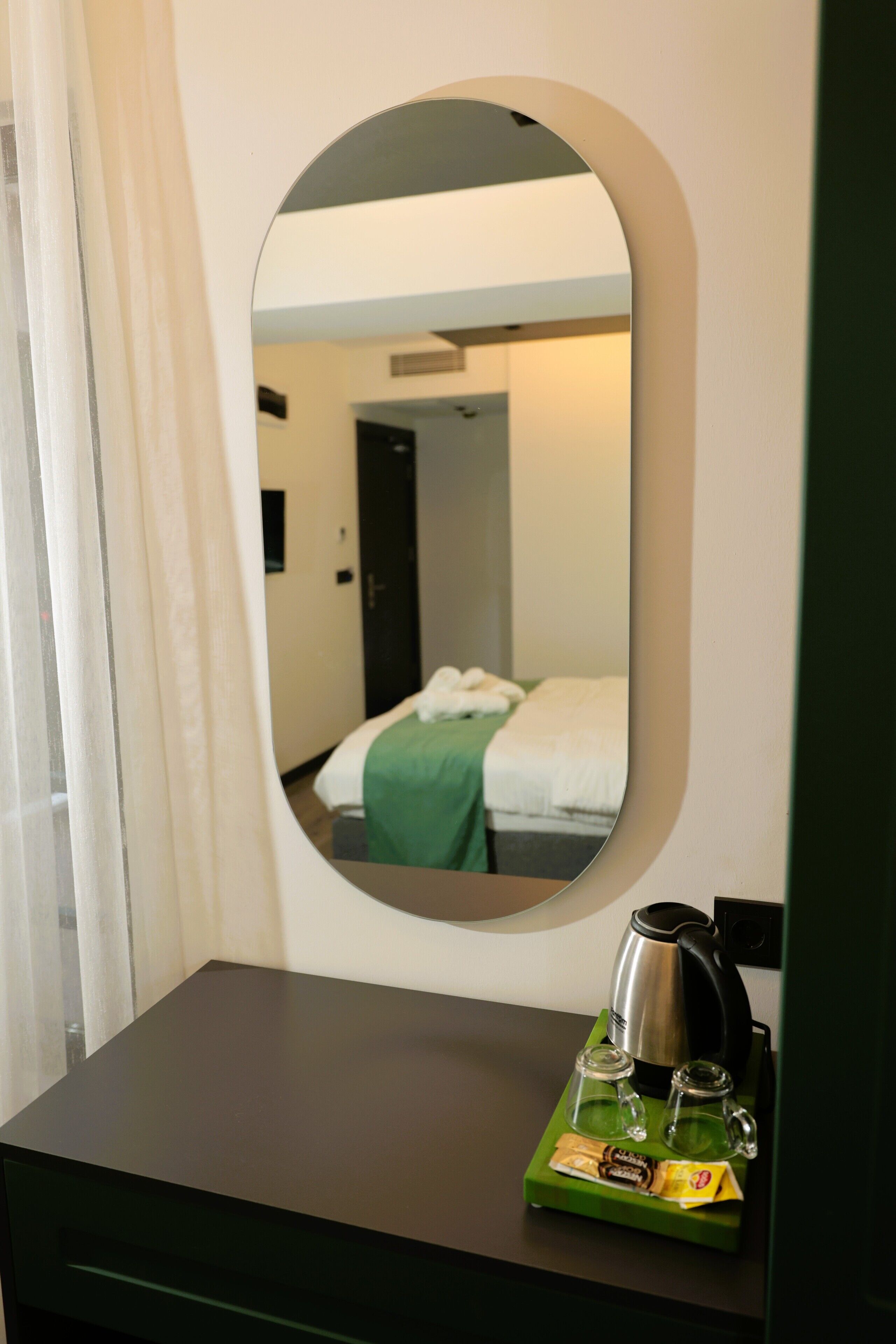 Habitación de hotel con cama, mesita de noche verde, hervidor y espejo que refleja la habitación.