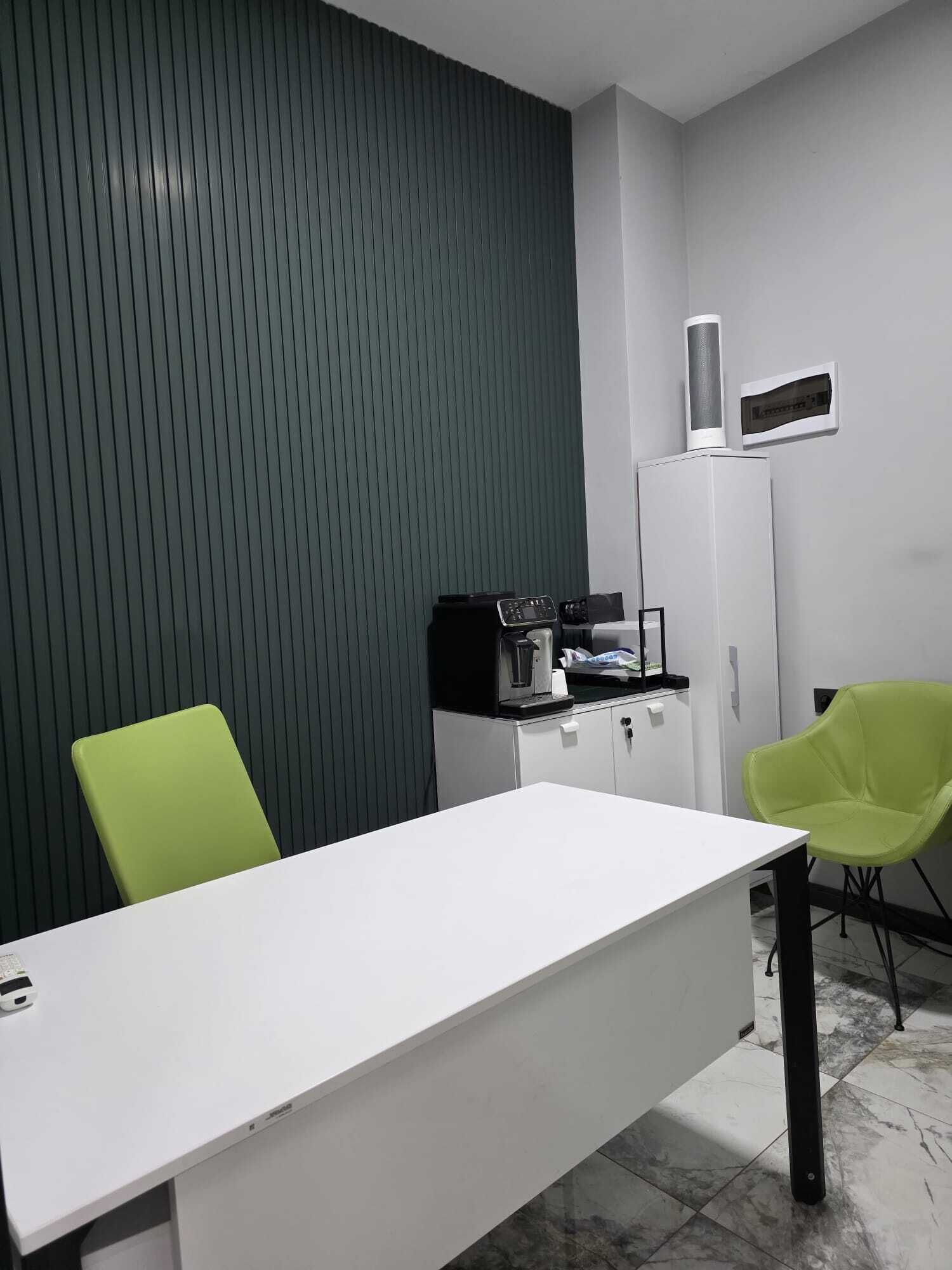 Une petite pièce moderne, avec un bureau blanc, deux chaises vertes, une machine à café noire et un meuble blanc.