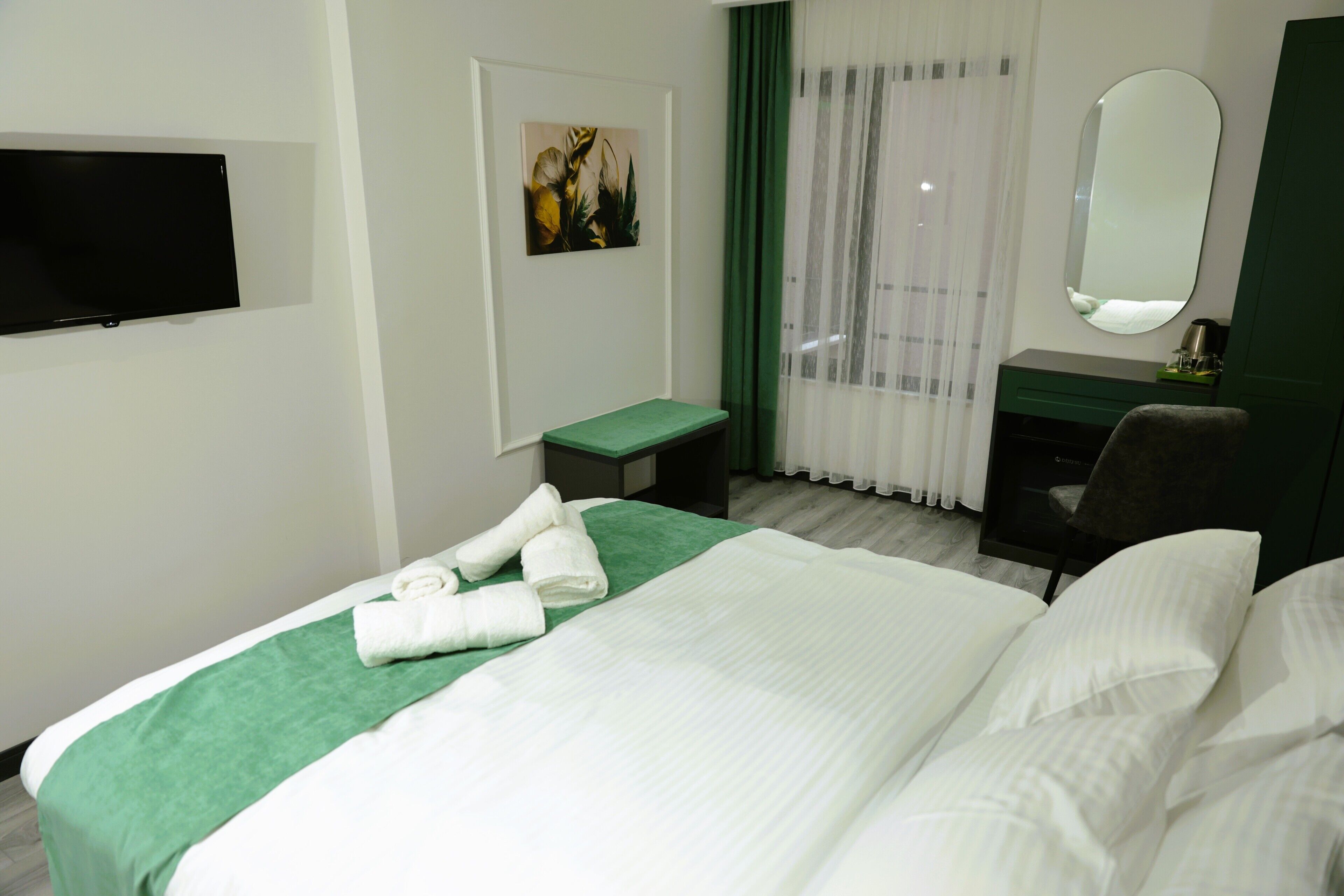 A hotel room with a bed, a TV mounted on the wall, a desk, a chair, and a window with curtains.