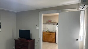 A room with a TV on a dresser, a door leading to a kitchen with wooden cabinets and a tiled backsplash. - Taminaka Condominium Comfortable II (Oranjestad)