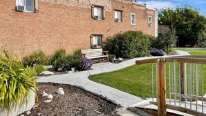 Edifici de maó amb un passeig pavimentat, una banca de fusta i una petita zona de jardí amb plantes i pedres.