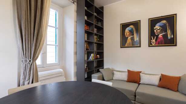 A modern living room with a dark wood dining table, a grey sofa with orange cushions, and a bookshelf filled with books and decorative items. Two paintings of women in blue headscarves are displayed on the wall.