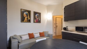 A modern living room with a grey sofa, a round table, and a kitchen area with dark cabinets and a microwave.