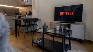 A modern living room with a flat-screen TV displaying 'NETFLIX', a glass coffee table, a sofa, and a kitchen area in the background. - Modern Apt. ideal for families/couples,10 mins from Thessaloniki's vibrant heart (Thessaloniki)