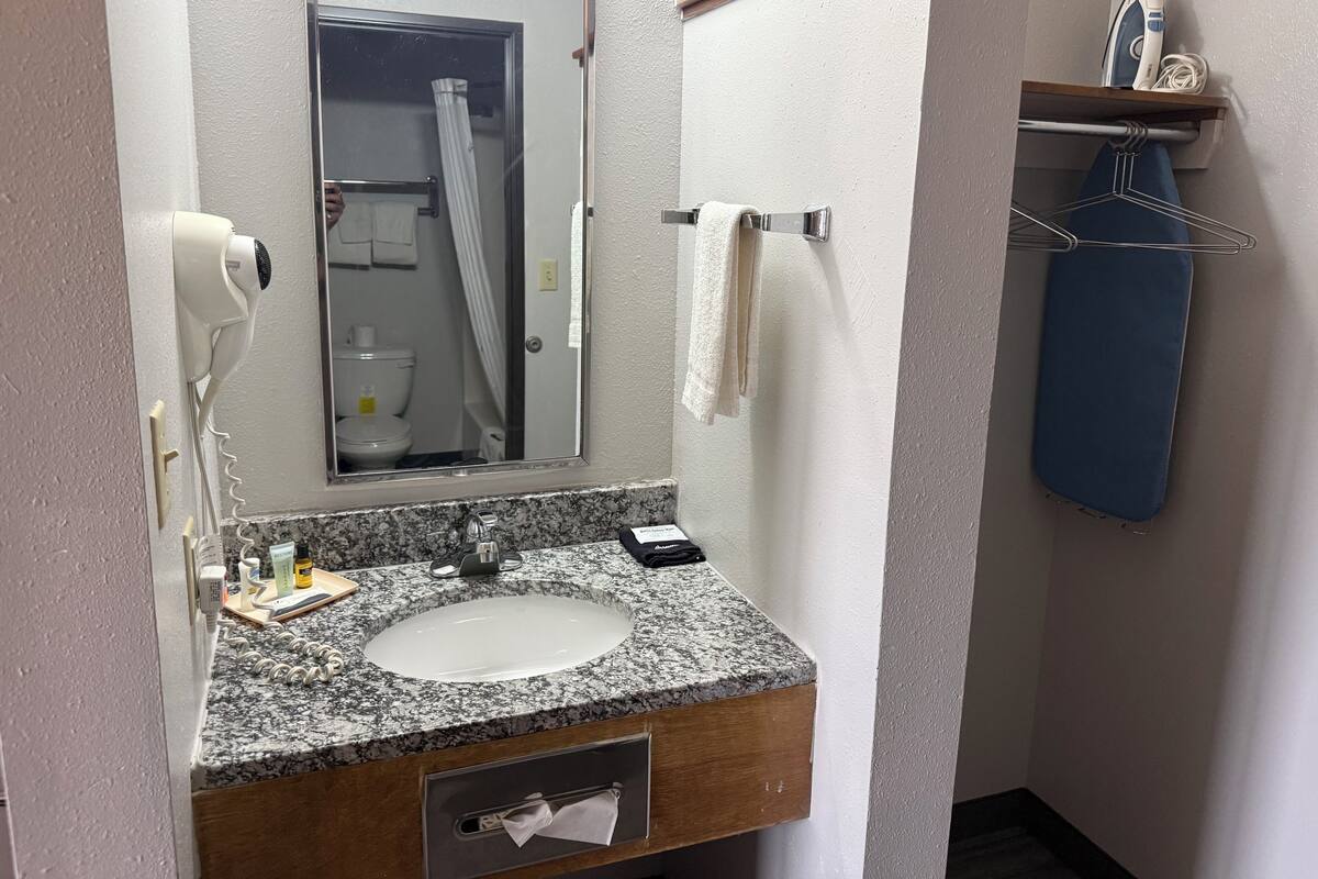 A bathroom with a granite countertop sink, a mirror, a hairdryer, a towel rack, and a shelf with an iron and a hanger.