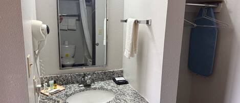 A bathroom with a granite countertop sink, a mirror, a hairdryer, a towel rack, and a shelf with an iron and a hanger.