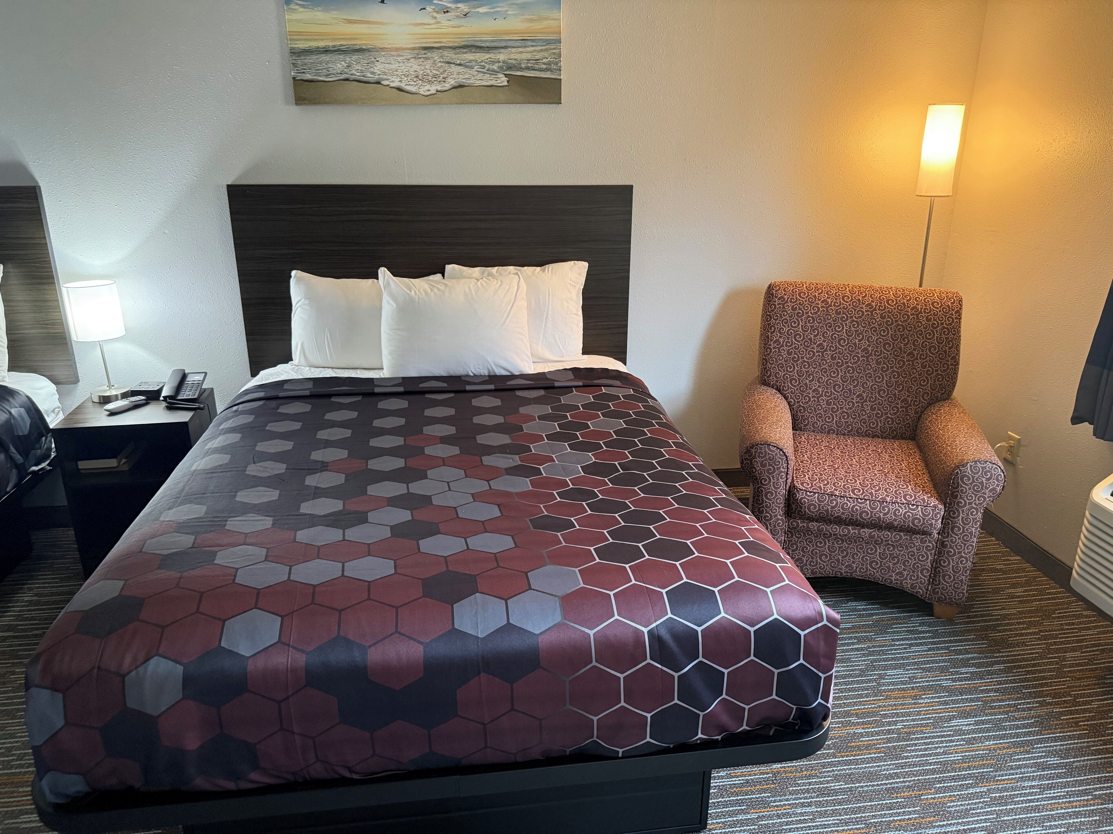 Quarto de hotel com uma cama, uma mesa de cabeceira com abajur, uma cadeira e um quadro na parede.