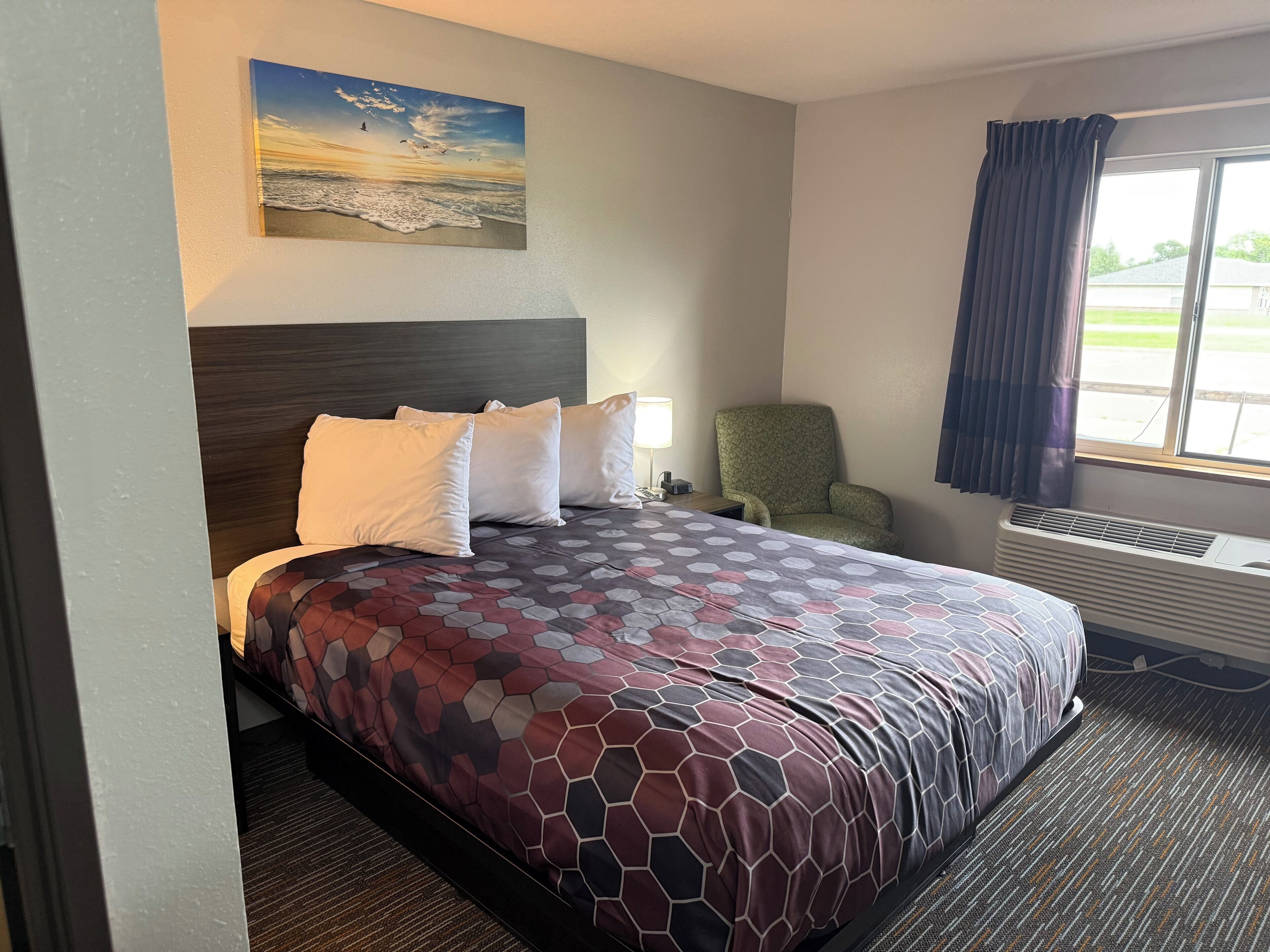 Ein Hotelzimmer mit Bett, Stuhl, Fenster mit Vorhängen, einem Wandbild und einem Heizkörper.