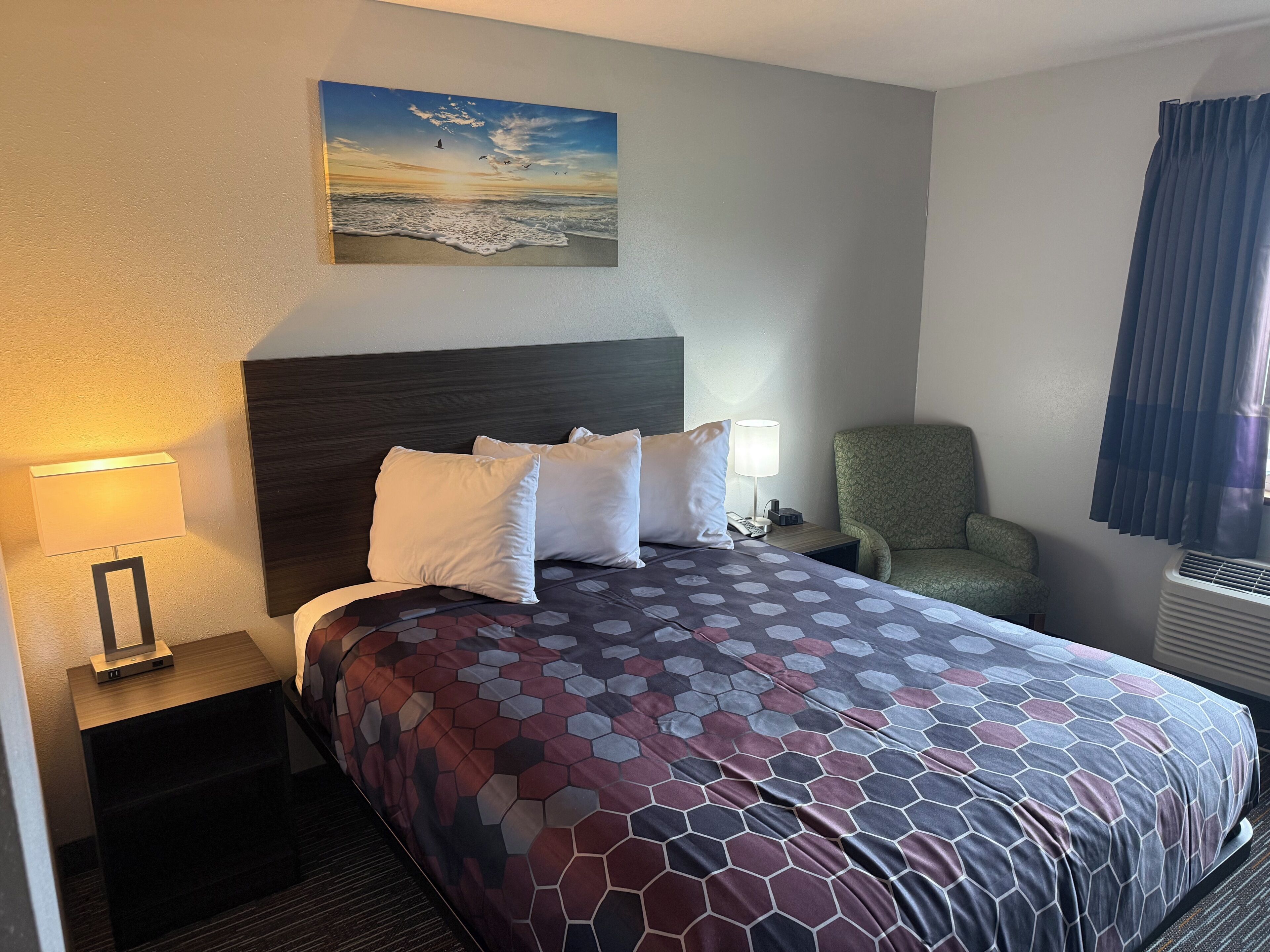 Een hotelkamer met een bed, nachtkastje, stoel, schilderij en een raam met gordijnen.