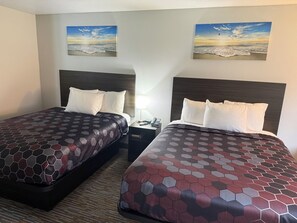 Two beds with hexagonal patterned bedding, a nightstand with a phone, and two wall-mounted paintings of a beach scene. - Country Haven Inn (Burlington)
