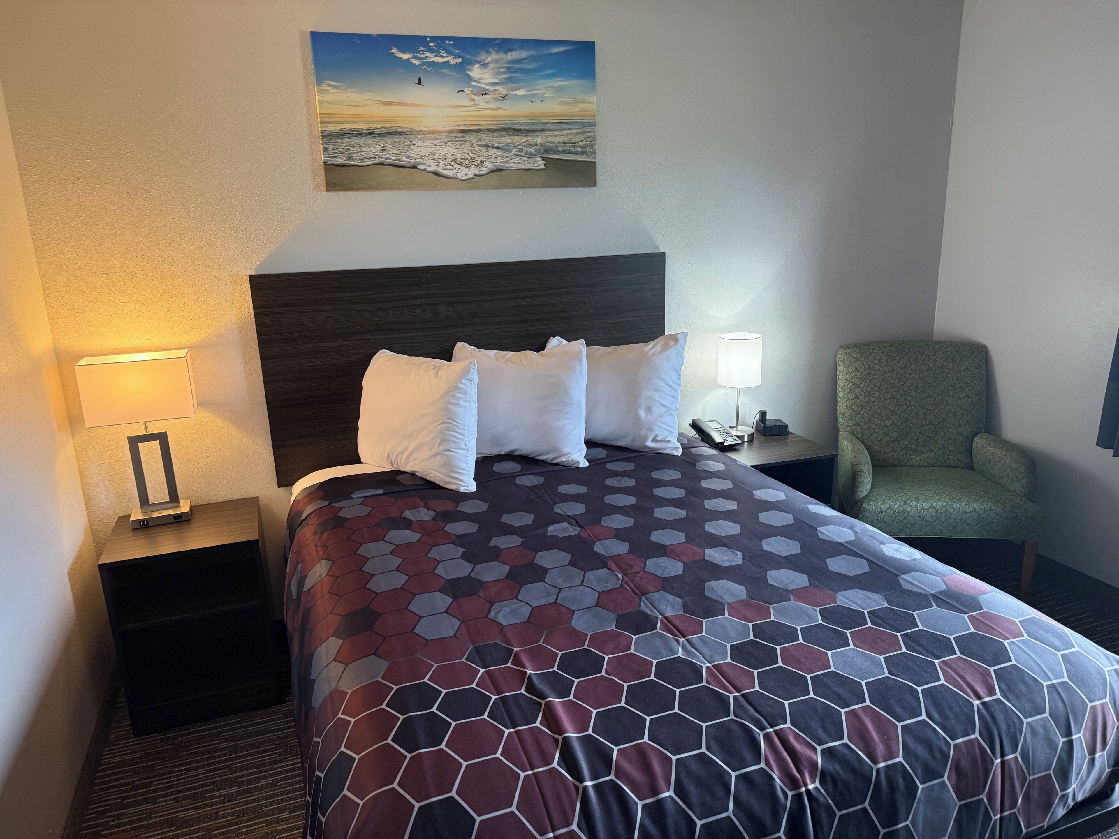 Une chambre d’hôtel avec un lit, une table de chevet avec une lampe, une chaise et un tableau représentant une scène de plage.