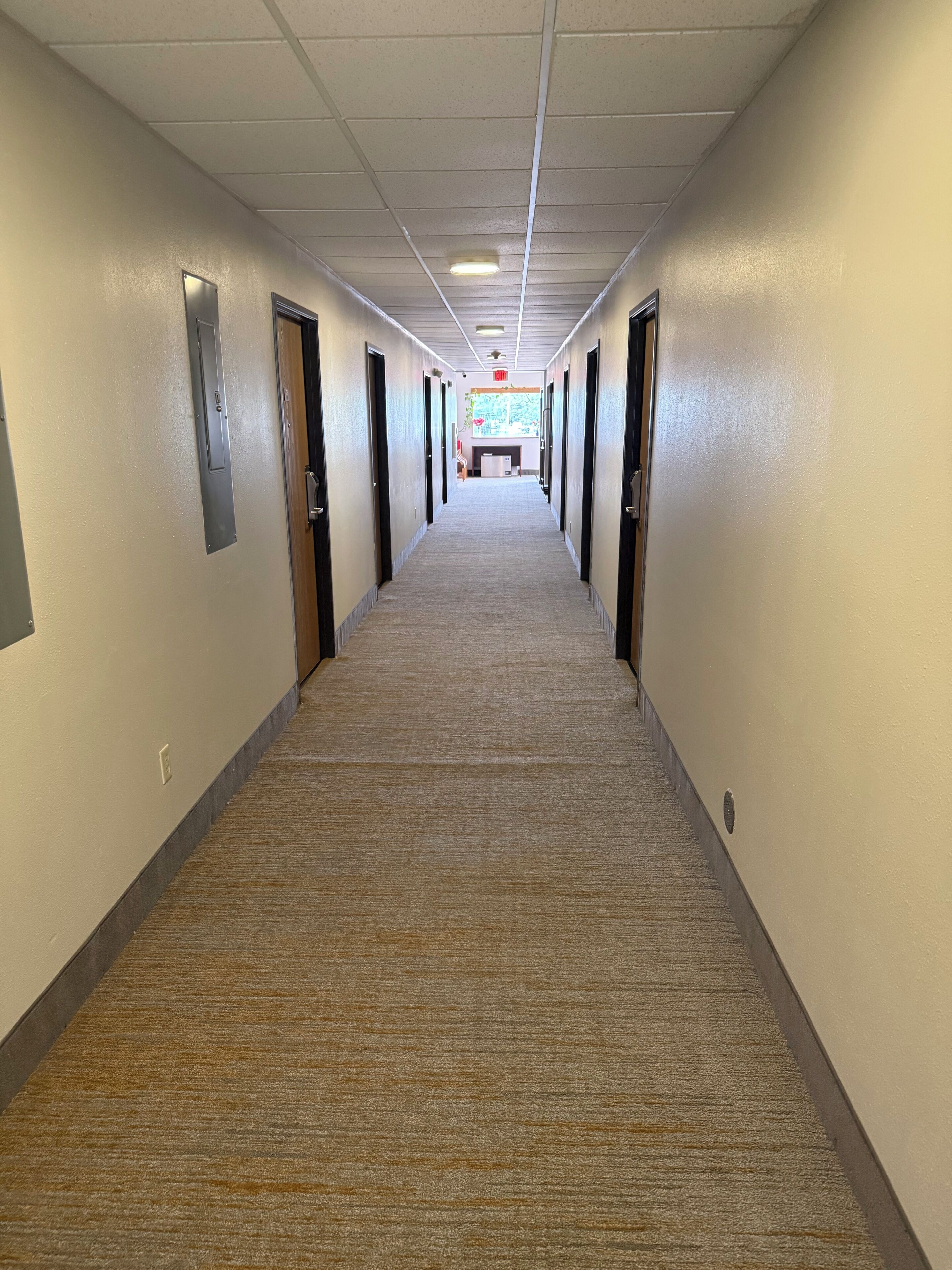 Un long couloir recouvert de moquette, avec des portes des deux côtés, qui mène à une pièce dotée d’une fenêtre et d’un évier.