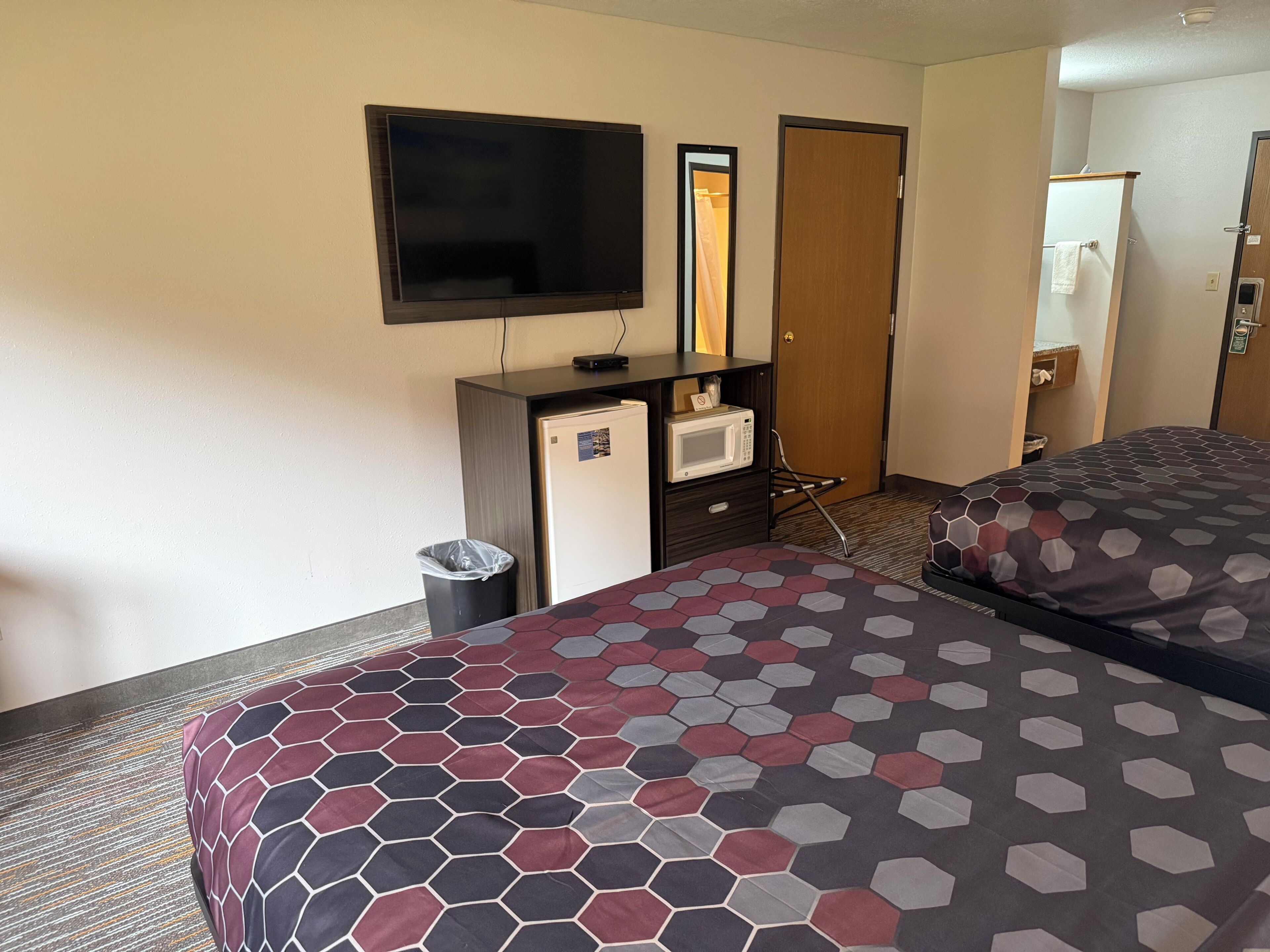 A hotel room with a bed, a TV, a desk with a microwave, a trash bin, and a door.