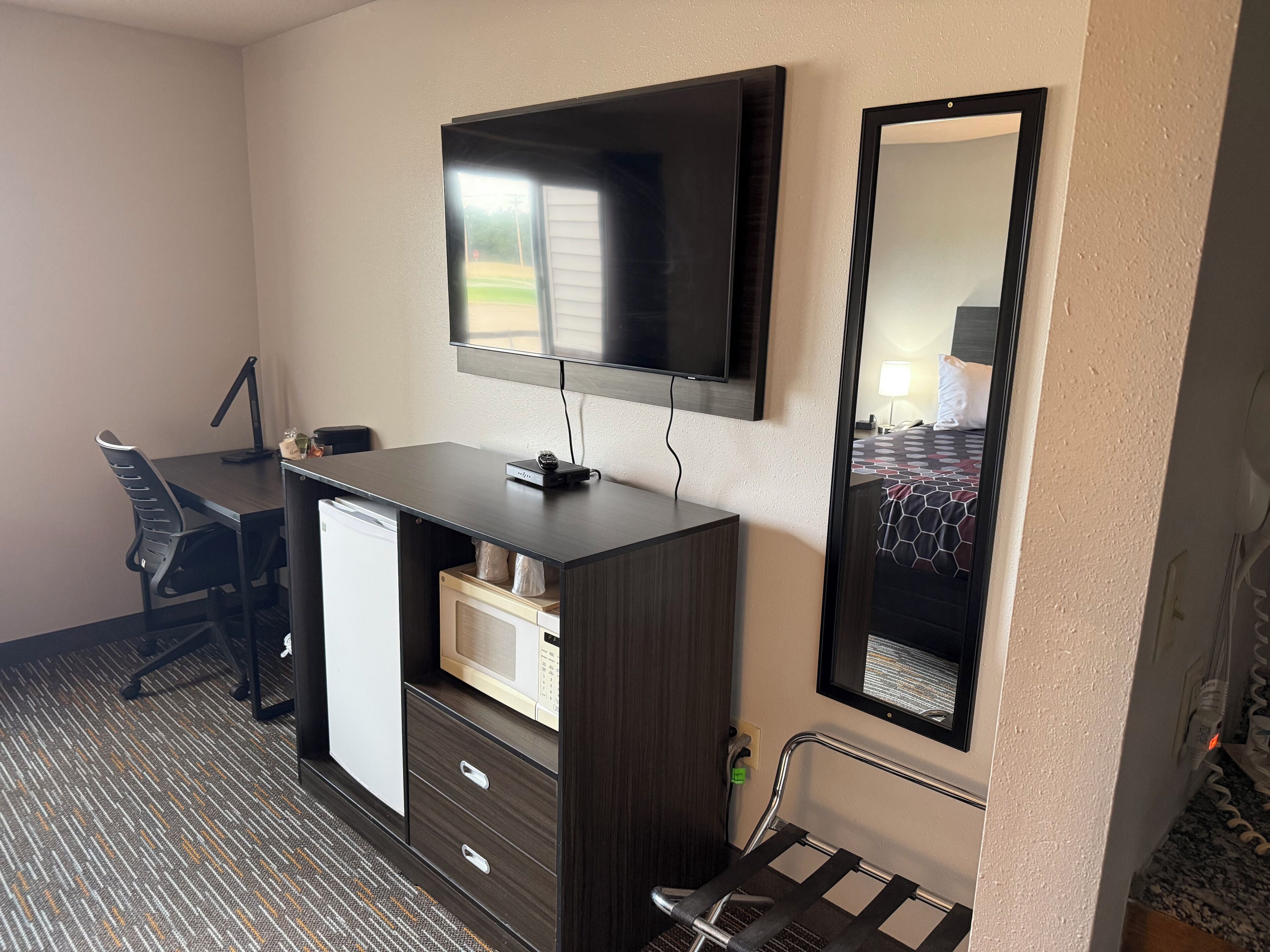 A hotel room with a desk, chair, TV, mirror, and a bed with a patterned bedspread.